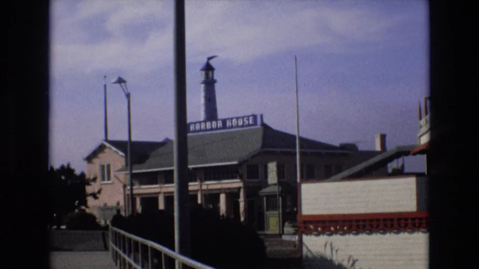 1973 8mm view of the Harbor House waterfront building on the Central Coast of California, with a lighthouse-style tower, railings, palm trees, and part of a neighboring structure.