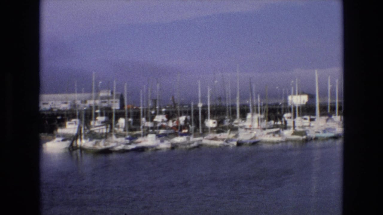 1973 Central Coast California 8mm frame showing a distant marina with sailboats, clustered masts, calm water, and a pale overcast sky.