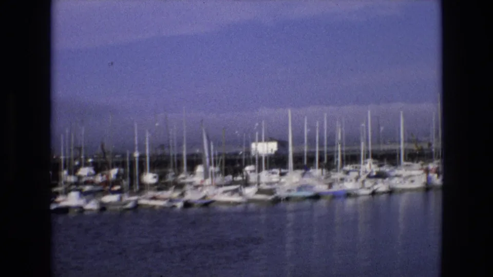 1973 Central Coast California 8mm frame with a broad view of moored boats, many vertical masts, still water, and low shoreline structures.