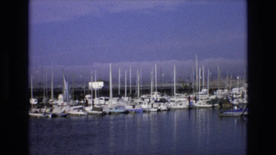 1973 Central Coast California 8mm frame showing a closer harbor section with moored sailboats, mast reflections, calm water, and a small white structure near the shore.