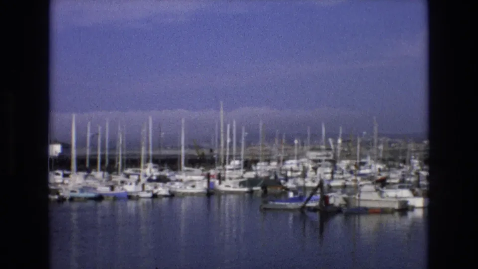 1973 Central Coast California 8mm frame showing a quiet marina scene with clustered sailboats, tall masts, smooth harbor water, and distant waterfront structures.