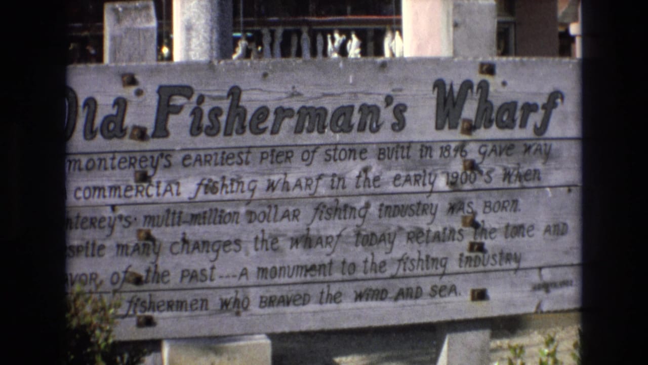 1973 8mm view of the Old Fisherman's Wharf sign, with weathered boards, handwritten lines about the wharf, and background railing partly visible above it.