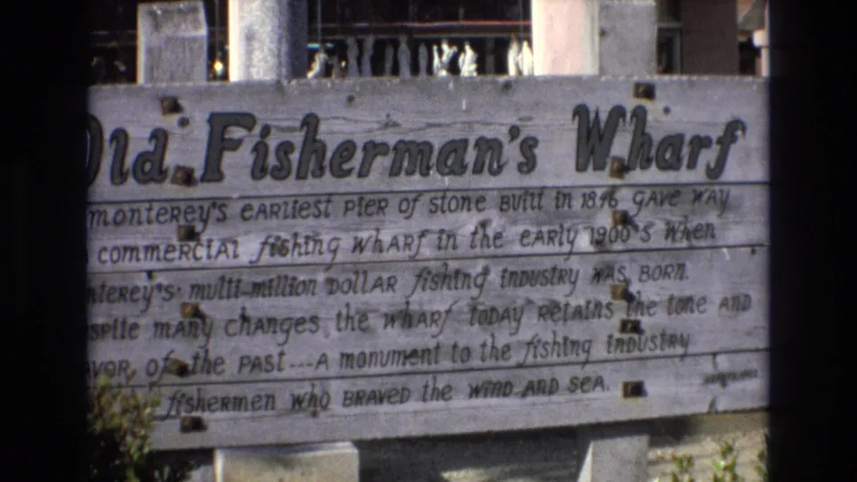 1973 8mm view of the Old Fisherman's Wharf sign, with weathered boards, handwritten lines about the wharf, and background railing partly visible above it.