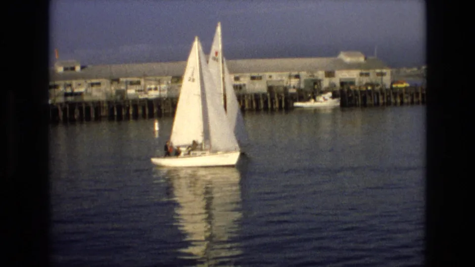 White sailboat carrying distant people on calm water near Old Fisherman's Wharf on the California coast, with dock pilings and waterfront buildings behind it.