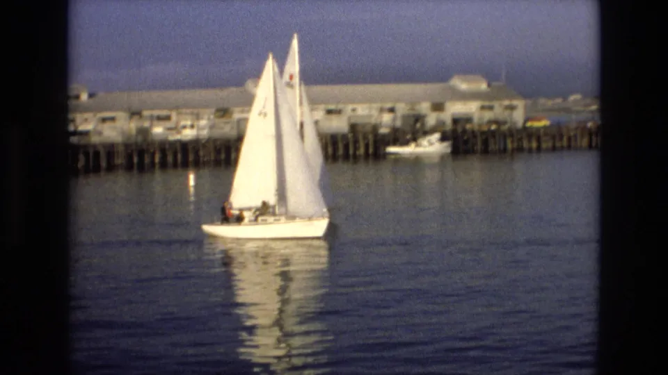 White sailboat with raised sails moves across calm harbor water near the California wharf, with distant waterfront buildings and dock structures in the background.