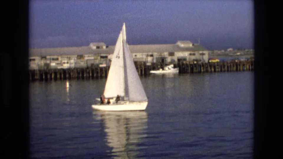 A white sailboat with passengers remains in view near the pier, with soft 8mm color and calm water emphasizing the home-movie harbor setting.