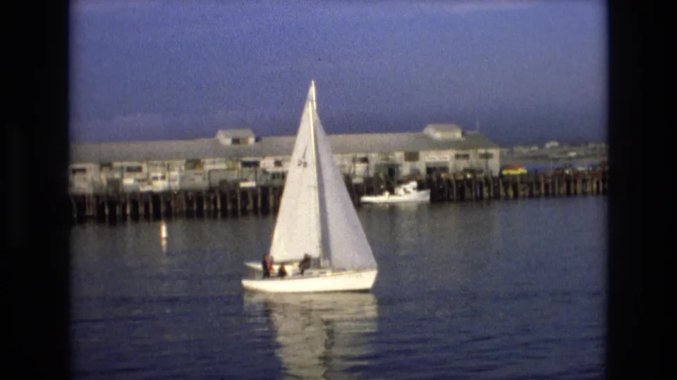 Near the end of the clip, the sailboat continues past the Central Coast California pier, with passengers visible as small figures and the sail reflected in the still water.