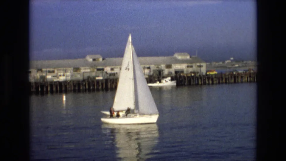 White sailboat marked with the number 29 moves across calm California coastal water near a wooden pier and low waterfront buildings in 1973 home-movie footage.