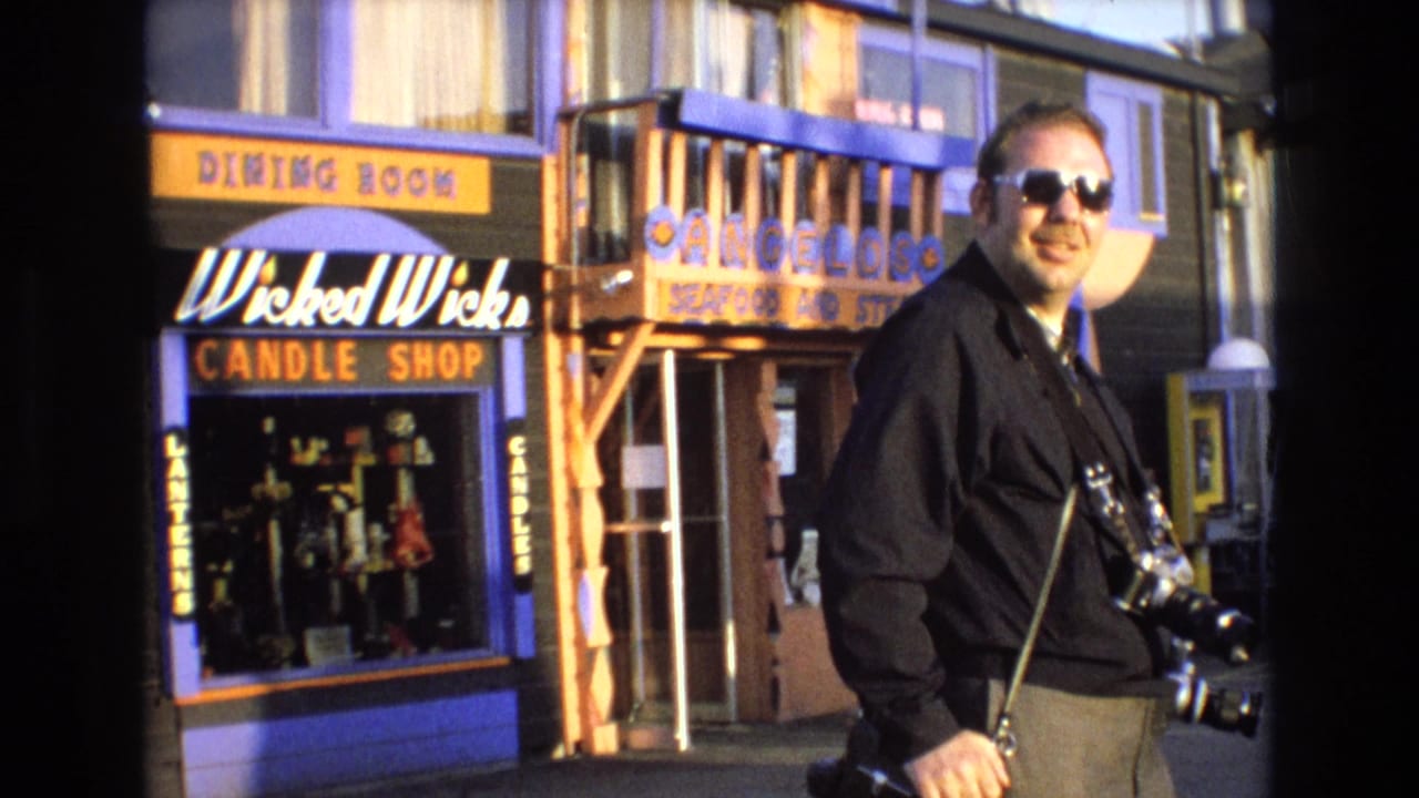Man in sunglasses and a dark shirt carries a camera while walking past colorful California coastal storefronts with visible candle and seafood shop signage.