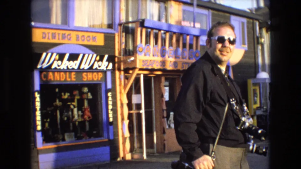 Man in sunglasses and a dark shirt carries a camera while walking past colorful California coastal storefronts with visible candle and seafood shop signage.