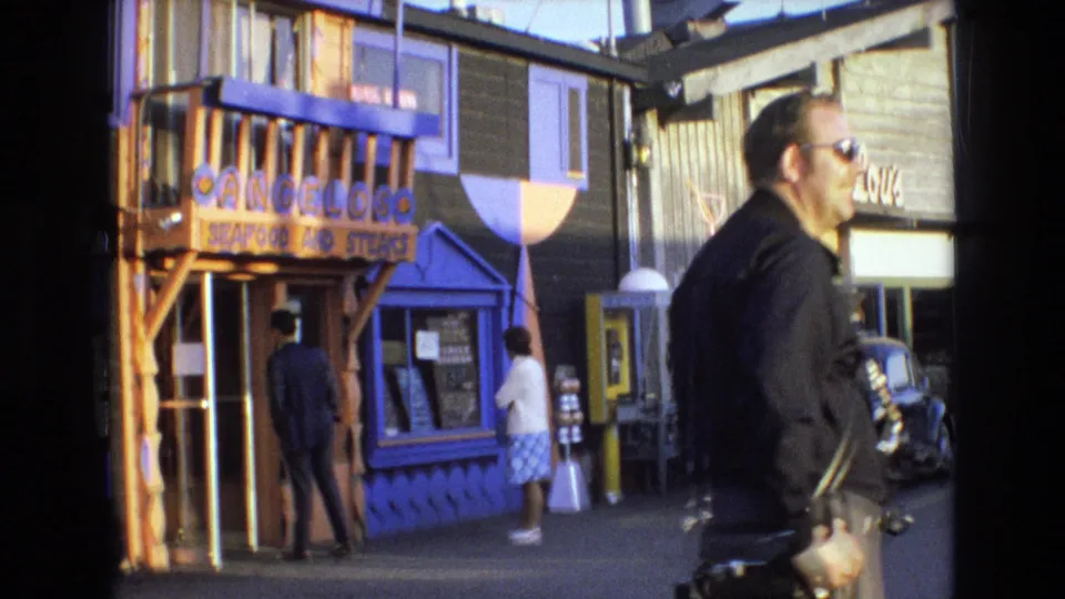 A man carrying a camera walks across the foreground while pedestrians stand near colorful waterfront shop entrances in 1973 California coastal home-movie footage.