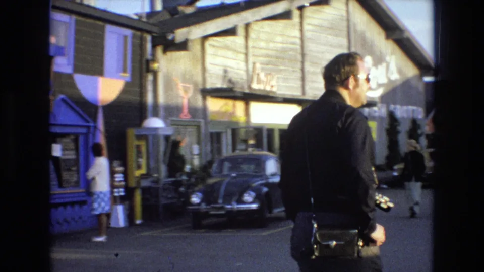 Man in sunglasses walks past waterfront shops with commercial signage and a black Volkswagen Beetle parked nearby in 1973 California coastal footage.