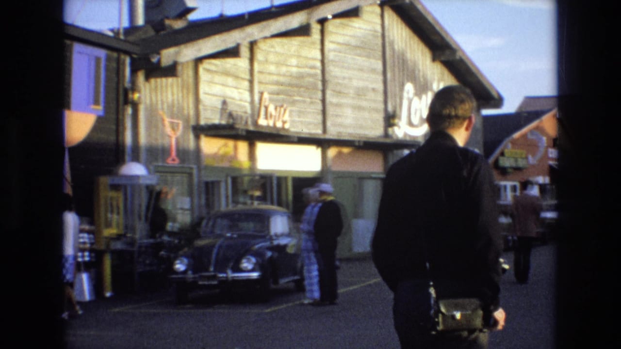 1973 Central Coast California home-movie frame showing a man from behind near Lou's Fish Grotto, with a black Volkswagen Beetle, pedestrians, and a wooden storefront visible.