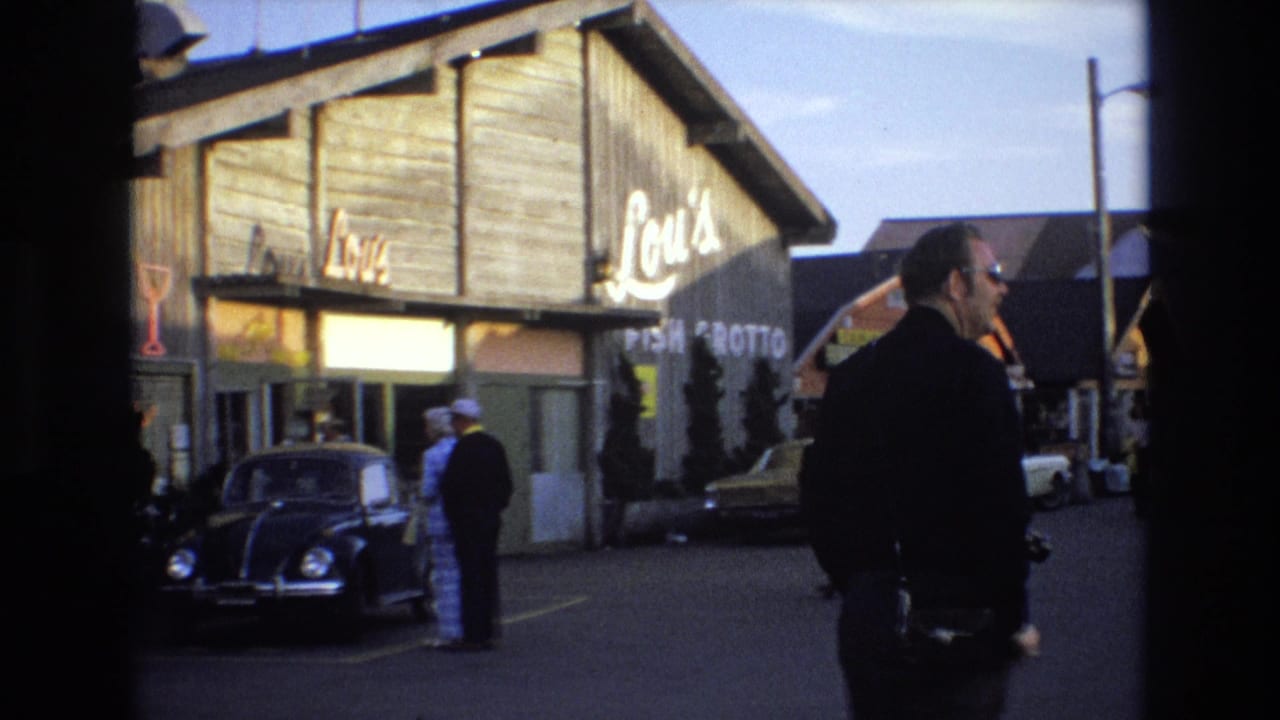 Man in sunglasses passes in front of Lou's Fish Grotto with a Beetle and pedestrians nearby.