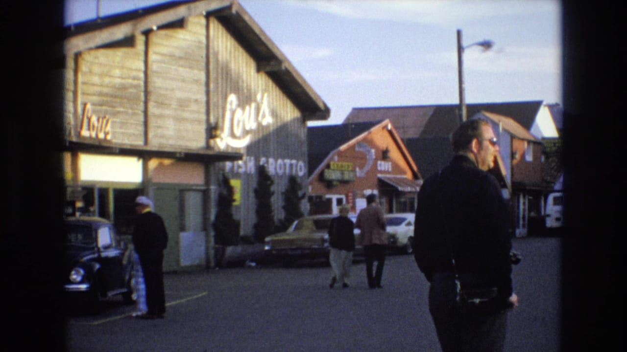 1973 Central Coast California home-movie frame showing Lou's Fish Grotto from the street, with pedestrians walking, a man in a dark jacket, a black Beetle, and neighboring commercial buildings.