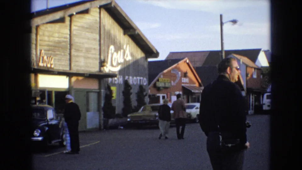 1973 Central Coast California home-movie frame showing Lou's Fish Grotto from the street, with pedestrians walking, a man in a dark jacket, a black Beetle, and neighboring commercial buildings.