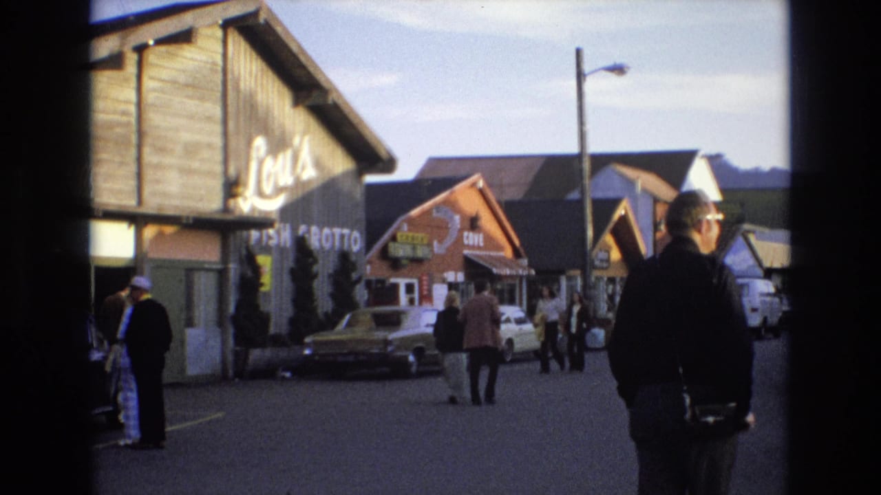 1973 Central Coast California street frame with Lou's Fish Grotto signage, a man at the right edge, pedestrians crossing the street area, parked cars, and neighboring storefronts.