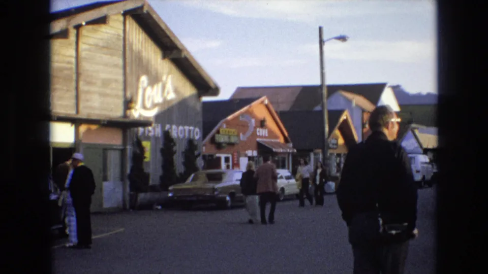 1973 Central Coast California street frame with Lou's Fish Grotto signage, a man at the right edge, pedestrians crossing the street area, parked cars, and neighboring storefronts.