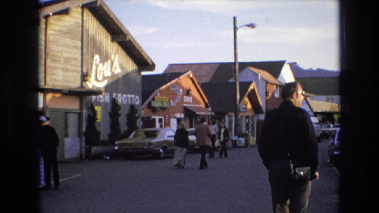 1973 Central Coast California 8mm frame showing a small commercial street with wooden storefronts, period cars, pedestrians, and visible Lou's Fish Grotto signage.
