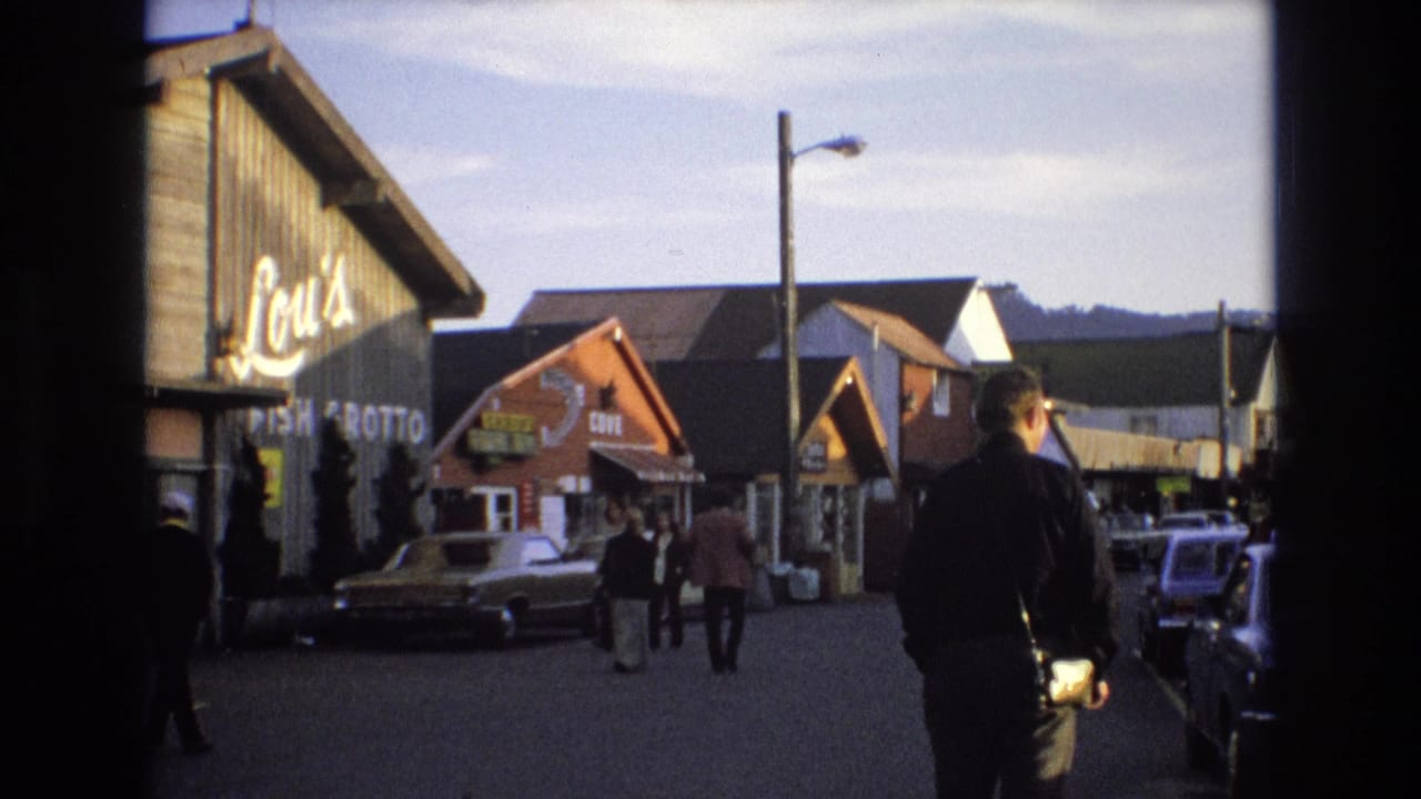 Man with a camera walks along a street lined with storefronts, parked cars, and pedestrians.