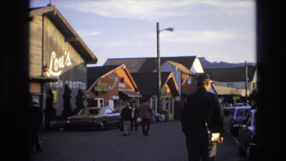1973 Central Coast California street view with a man carrying a camera, pedestrians, period cars, and the Lou's Fish Grotto storefront visible in the background.