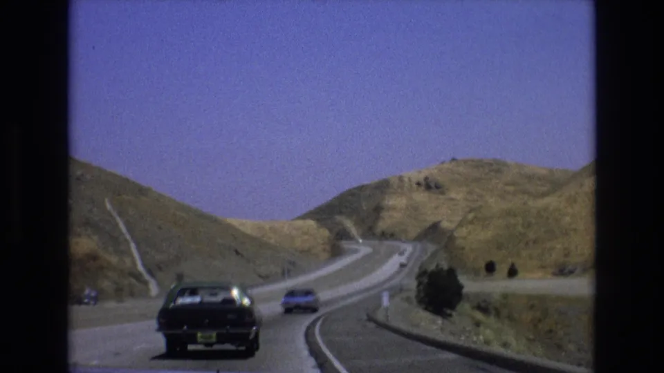 1973 Central Coast California 8mm frame of older cars driving along a winding highway through open dry hillside terrain.