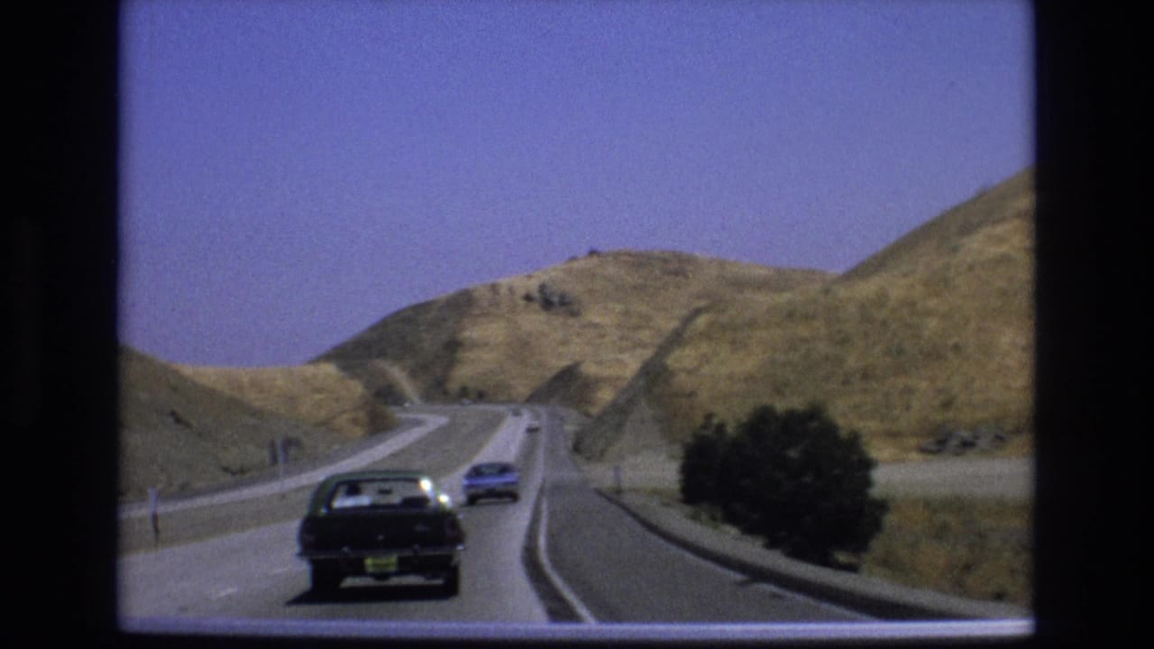 Cars drive ahead on a highway bordered by golden hills and shrubs.