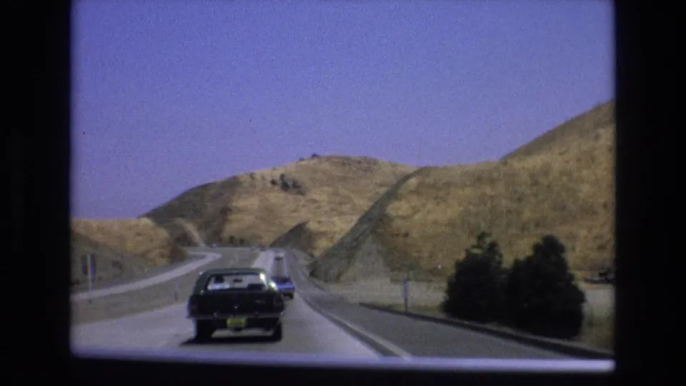 Rear view of a black sedan on a winding California road, with a blue car ahead and dry hillside terrain filling the background.