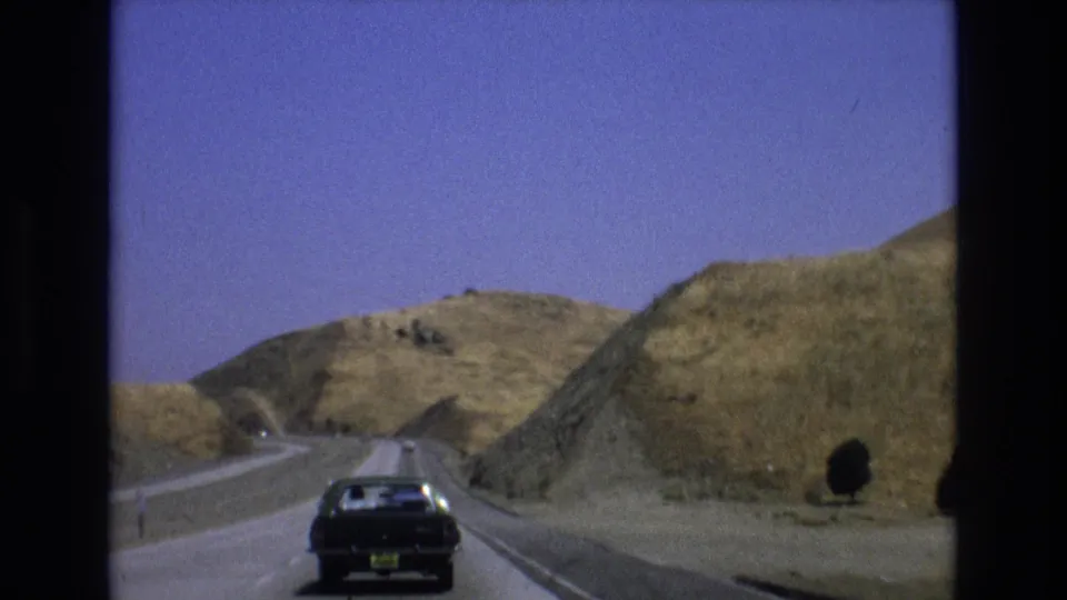 A black sedan travels on a curving highway through dry Central Coast California hills, framed by visible 8mm film borders and color-shifted sky.