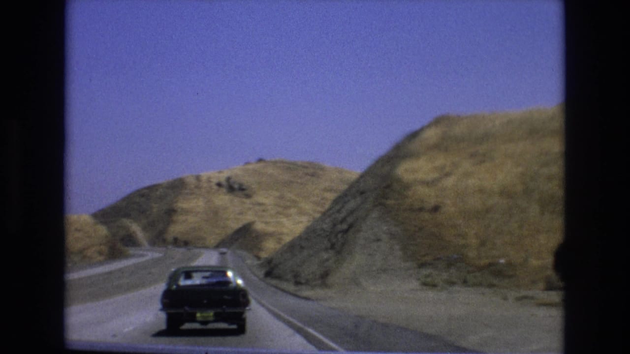 A dark period passenger car travels away from the camera on a sunlit Central Coast California road bordered by dry golden hills, consistent with the 1973 8mm home-movie metadata.