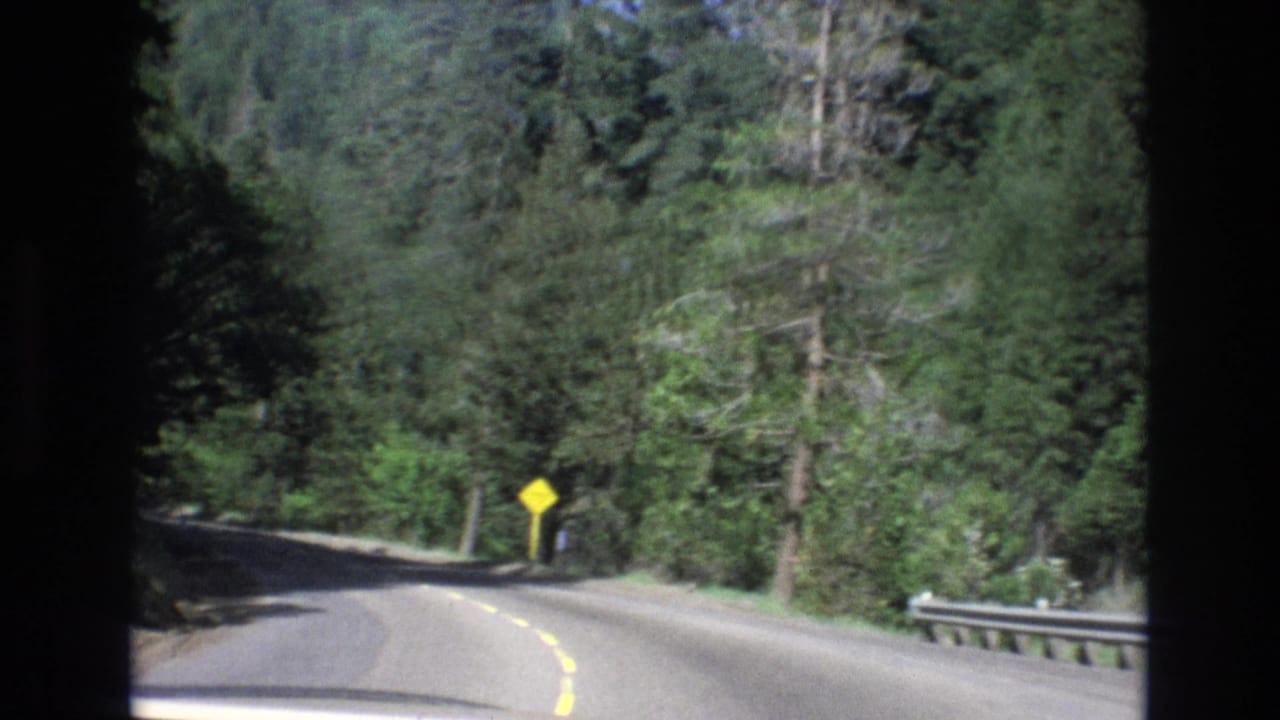 1973 8mm frame showing a two-lane road bending through dense Central Coast California forest, filmed from a car window with a yellow warning sign near the shoulder.