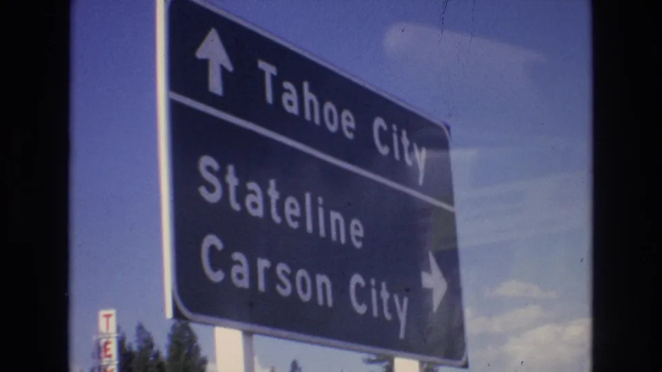 Blue roadside guide sign with arrows for Tahoe City, Stateline, and Carson City, seen in soft 8mm color with sky and trees behind it.