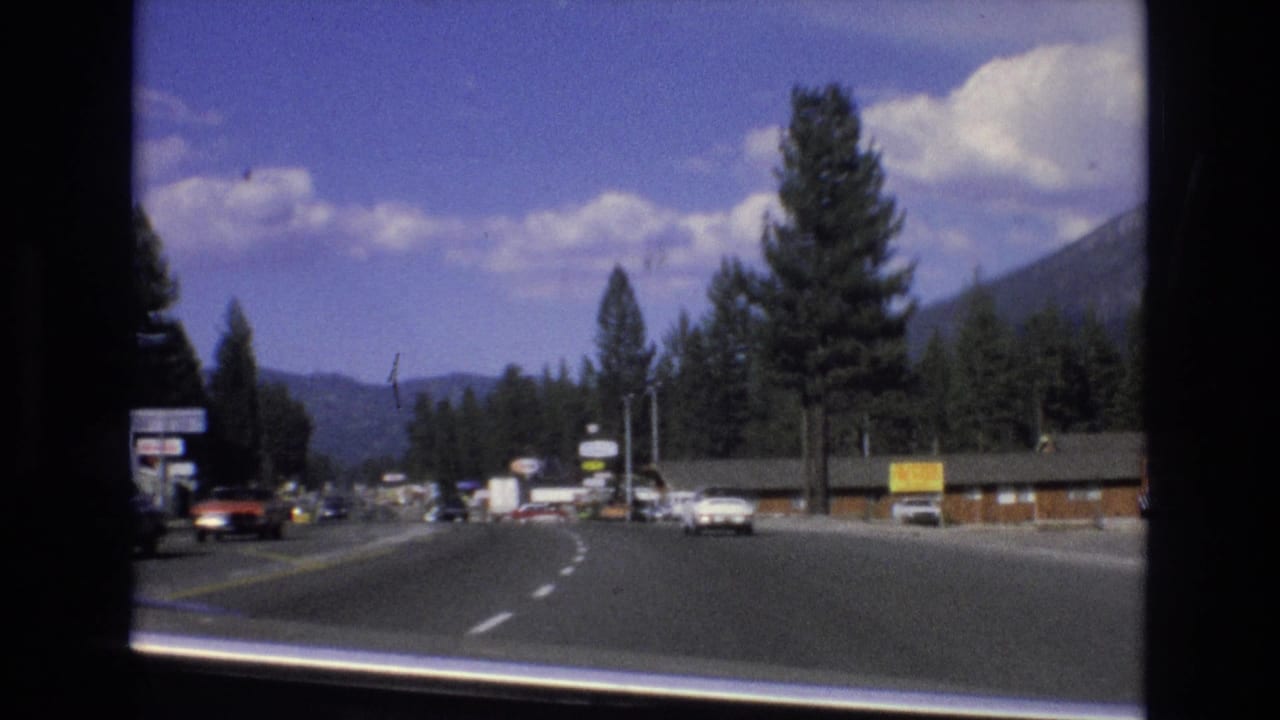 Moving-car view along a broad pine-lined roadway with period cars, roadside businesses, distant mountains, and dark window or film-frame borders.