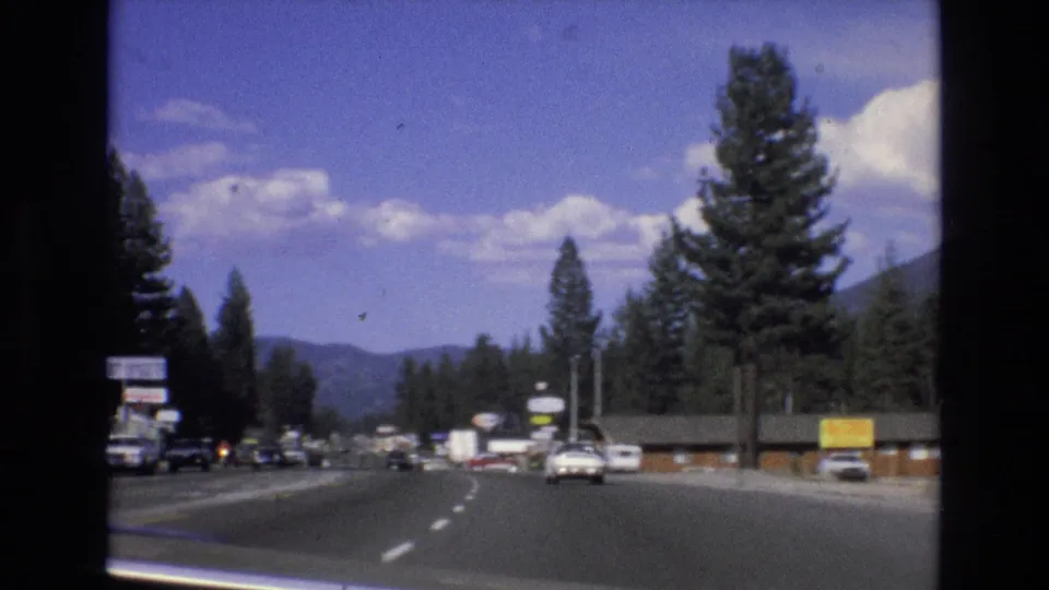 Soft 8mm road-travel frame showing a forested commercial corridor with traffic, roadside signs, low buildings, and mountain scenery ahead.