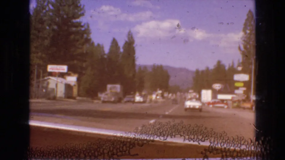Color-shifted 8mm frame of traffic on a forested roadway, with roadside signs, low commercial buildings, pine trees, and mountains visible beyond.