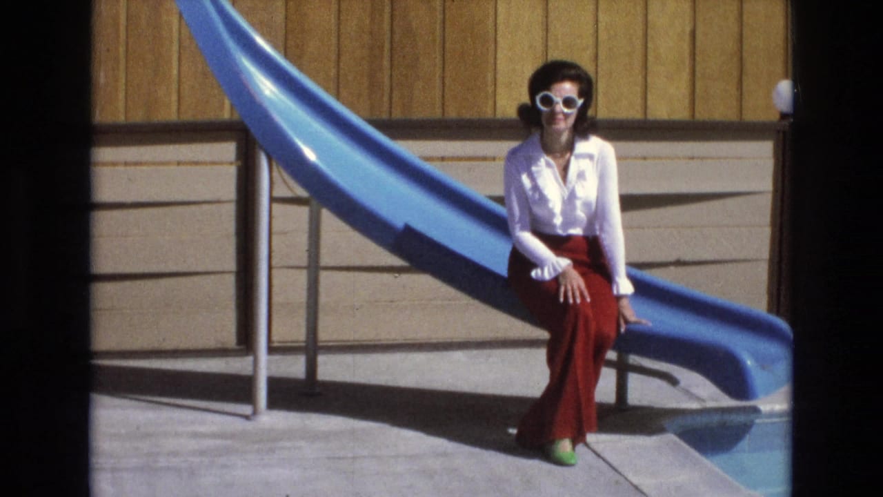 In 1973 Central Coast California home-movie footage, a woman in a white blouse and red pants sits at the base of a blue water slide by a pool.
