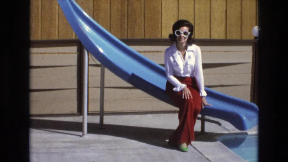 In 1973 Central Coast California home-movie footage, a woman in a white blouse and red pants sits at the base of a blue water slide by a pool.