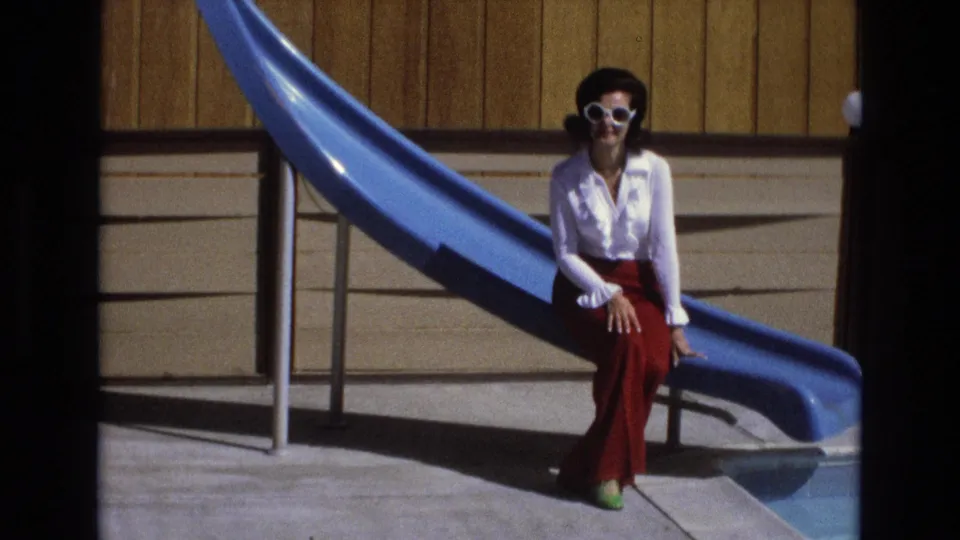 A woman wearing white-rimmed sunglasses sits near a blue water slide and pool edge, framed against a wood-paneled wall in 1973 California footage.