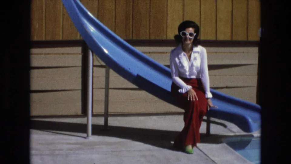 The woman sits casually in front of the blue slide, with red flared pants and green shoes visible near the pool edge in the 1970s home-movie frame.