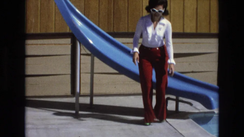 The woman has risen to stand beside the blue water slide, wearing a white blouse, red pants, white sunglasses, and green shoes near the pool.