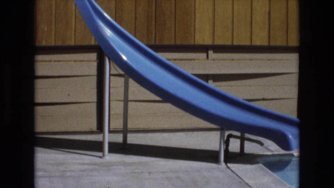 1973 8mm frame from Central Coast California showing a bright blue plastic pool slide, metal supports, concrete deck, pool edge, and wood-paneled wall in strong sunlight.