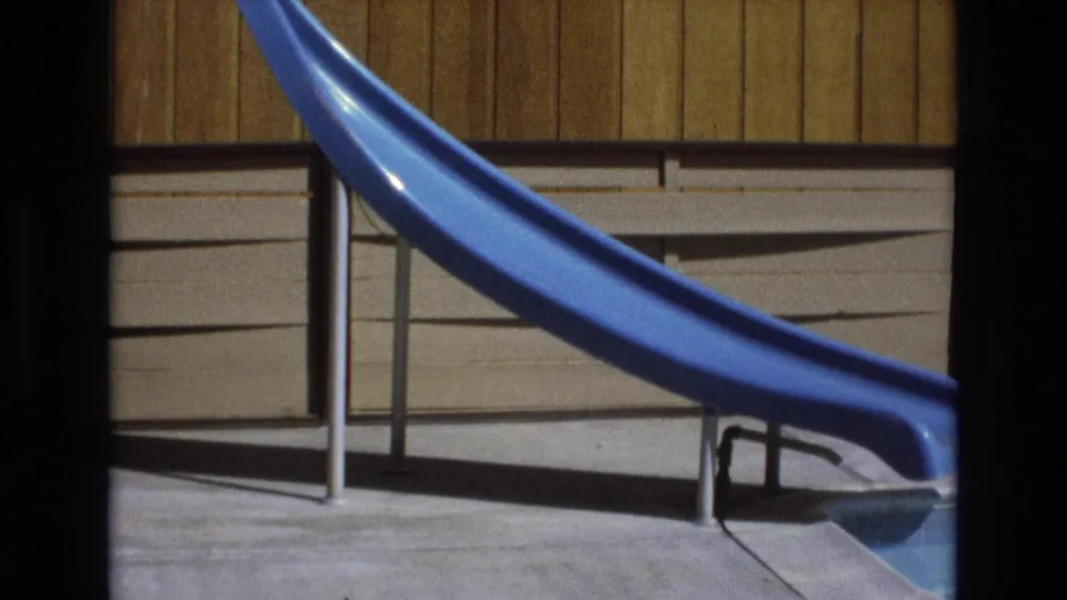 1973 8mm home-movie frame showing the curved blue pool slide more tightly framed, with upright metal supports, pool deck concrete, wall siding, and visible film grain.