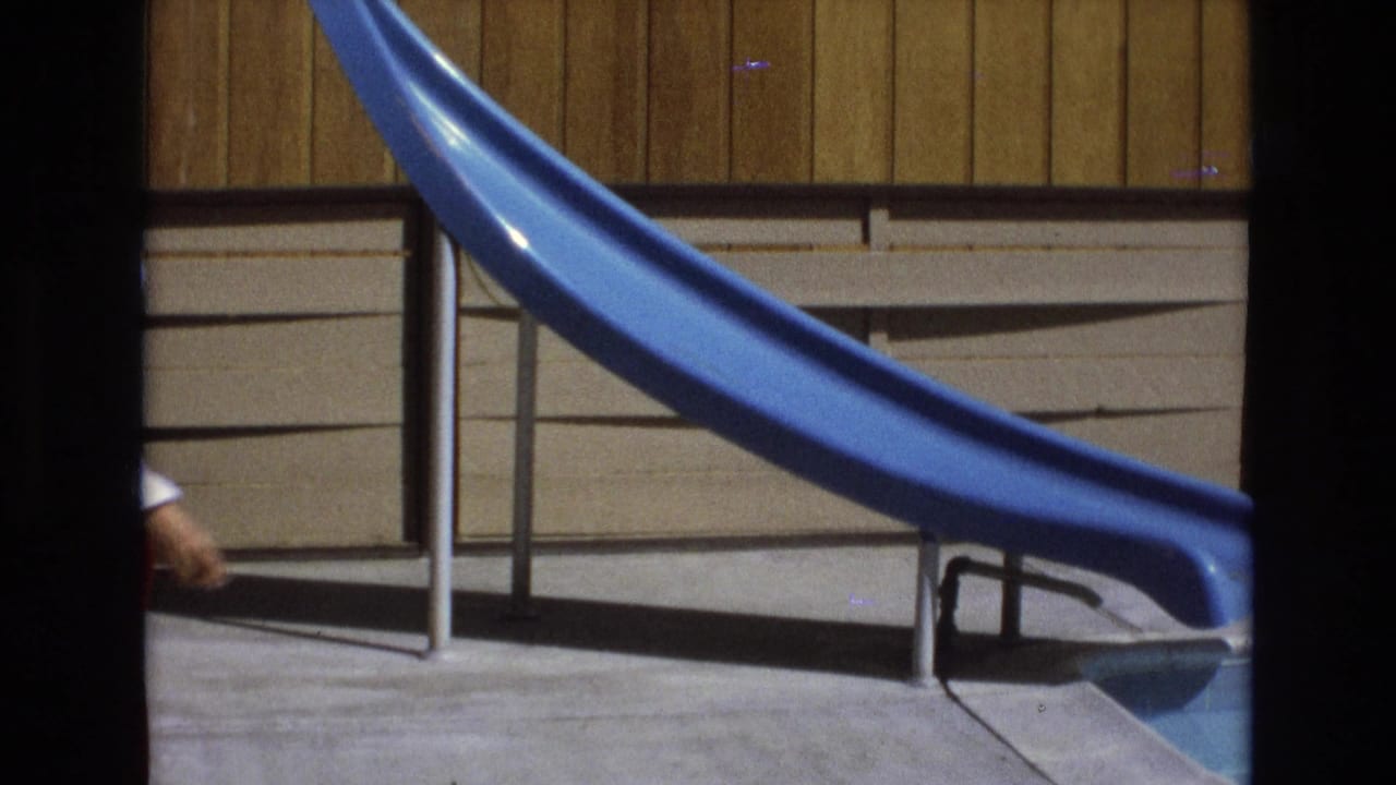A 1973 Central Coast California 8mm frame shows a blue pool slide, metal railing, wood-paneled wall, and concrete deck before the woman enters the center of view.