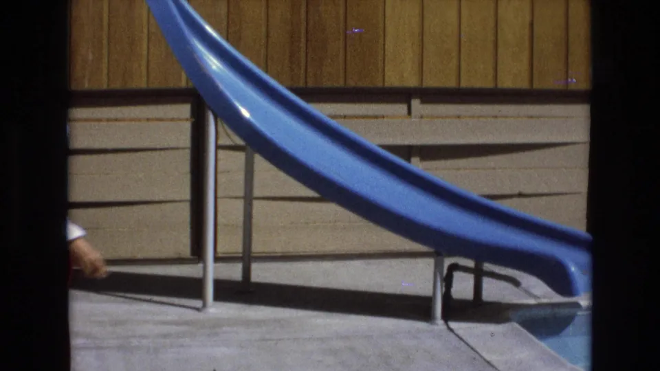 A 1973 Central Coast California 8mm frame shows a blue pool slide, metal railing, wood-paneled wall, and concrete deck before the woman enters the center of view.