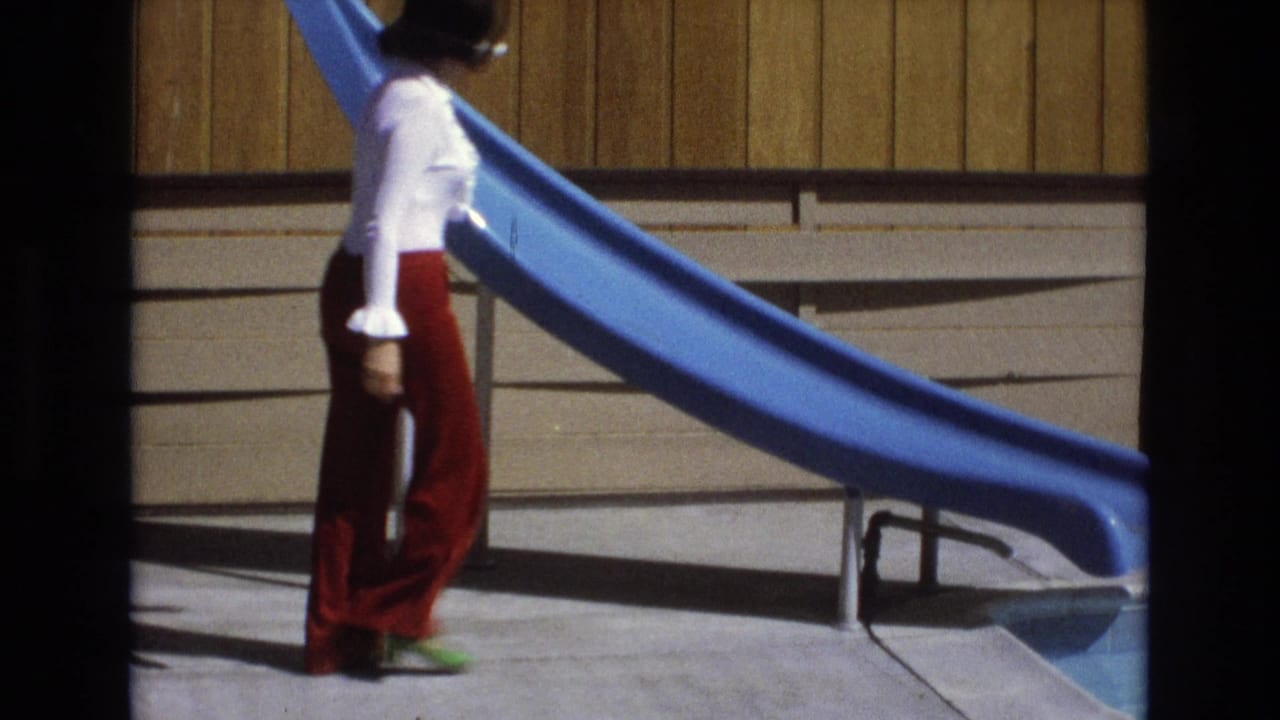 A woman in a white blouse, red flared pants, white sunglasses, and green shoes walks past a blue pool slide in the 1973 Central Coast California home-movie frame.