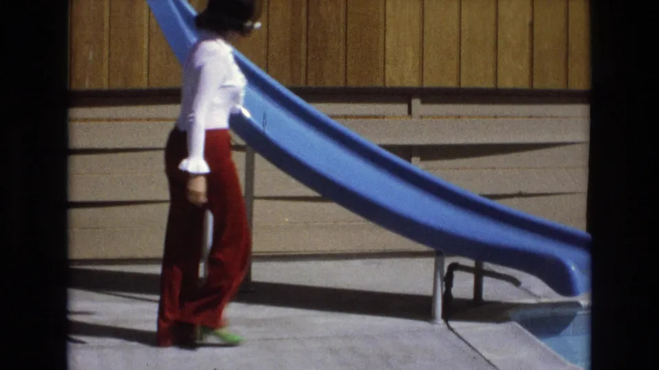 A woman in a white blouse, red flared pants, white sunglasses, and green shoes walks past a blue pool slide in the 1973 Central Coast California home-movie frame.