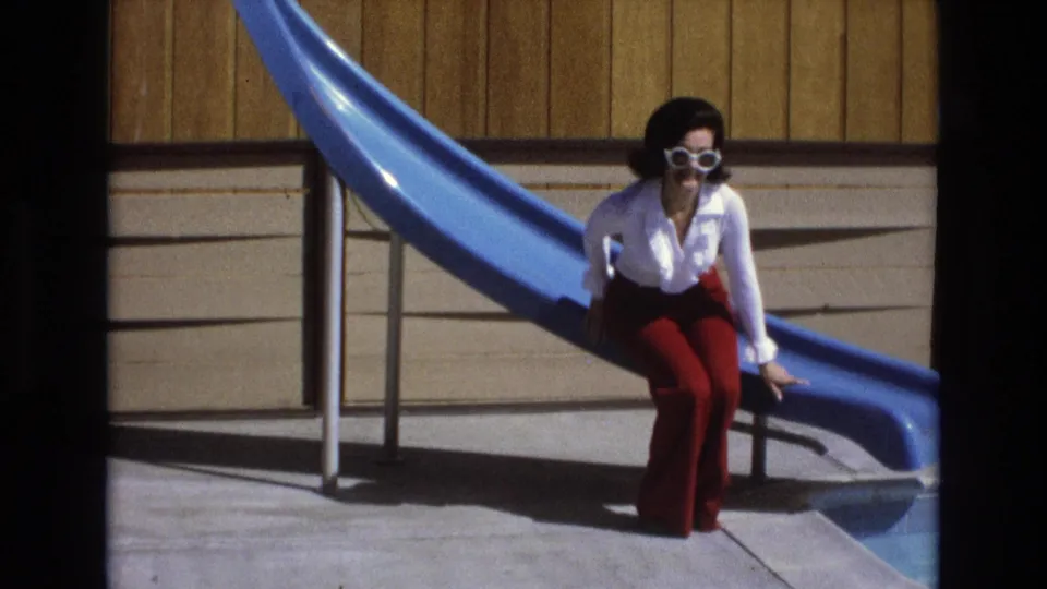 The woman sits at the edge of the blue pool slide, facing the camera in white sunglasses, a white blouse, and red pants against the wood-paneled outdoor wall.