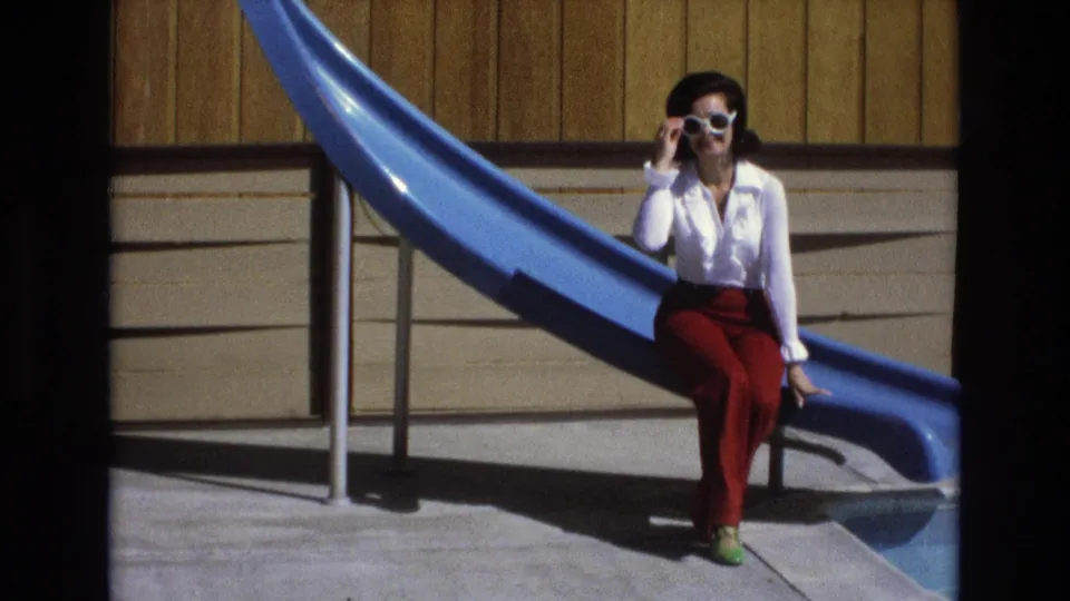 In the closing frame, the woman sits on the blue pool slide and adjusts or touches her white sunglasses while posing toward the camera in bright sunlight.