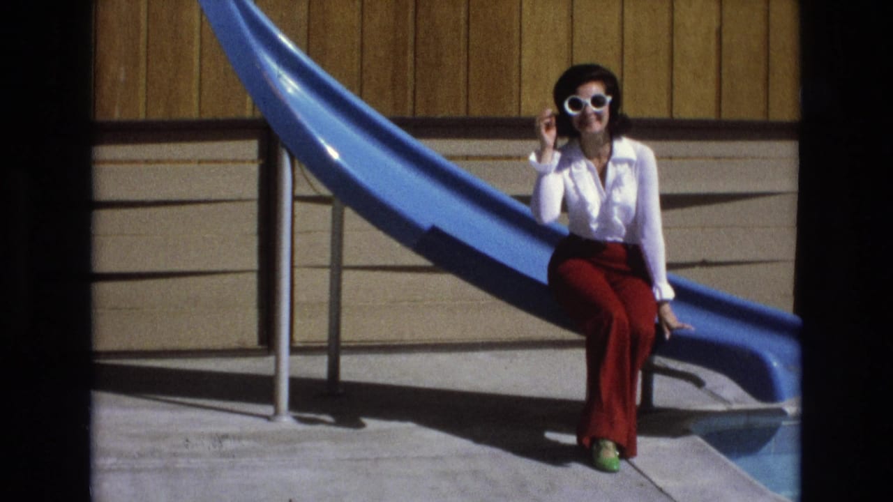 A woman in a white blouse and red pants sits beside a blue pool slide in 1973 Central Coast California home-movie footage, lifting one hand to her white sunglasses.