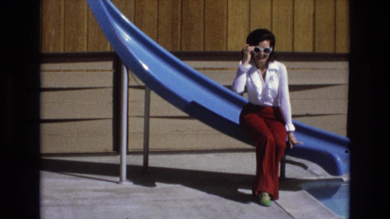 The seated woman remains beside the blue pool slide, holding her white sunglasses while facing the camera in bright daylight.